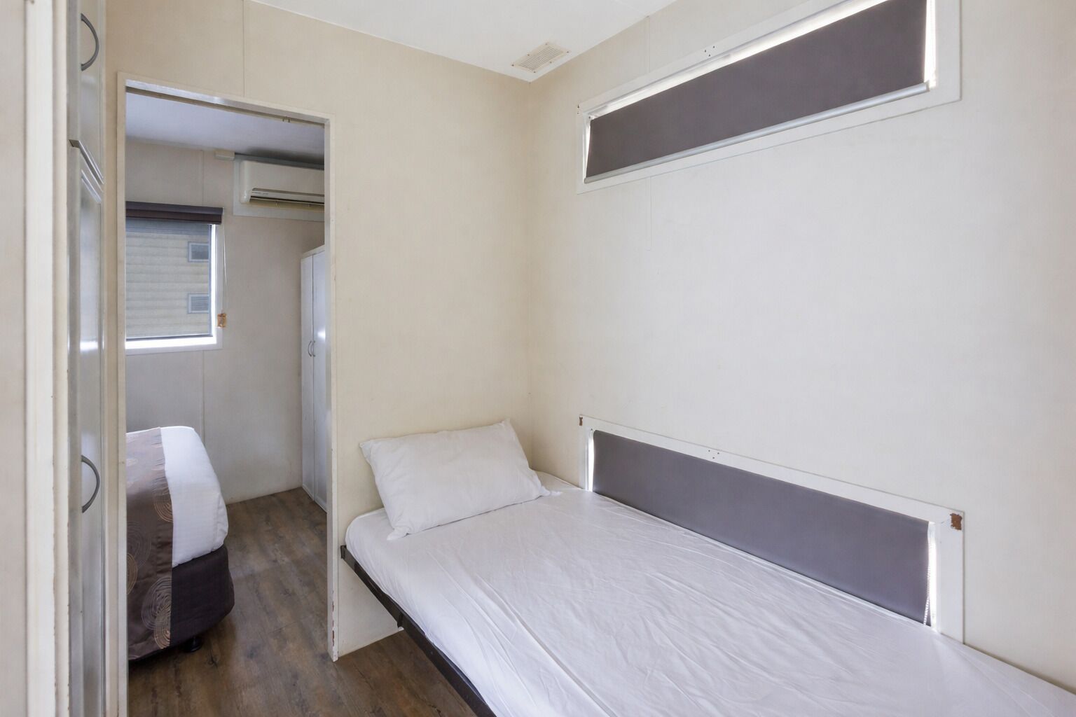 A small, single-bed room with a window, a wardrobe, and a door leading to another room.