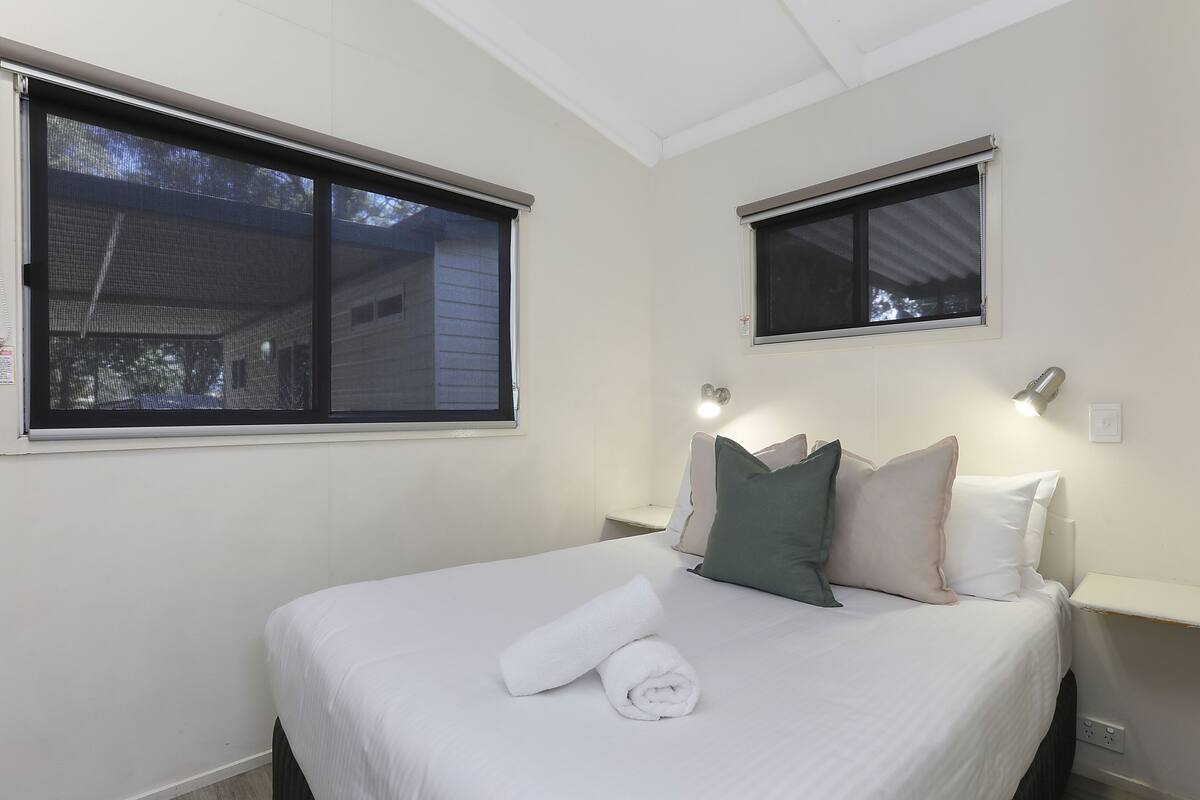 Une chambre d’hôtel avec un lit bien fait, deux fenêtres et une table de chevet avec une lampe.