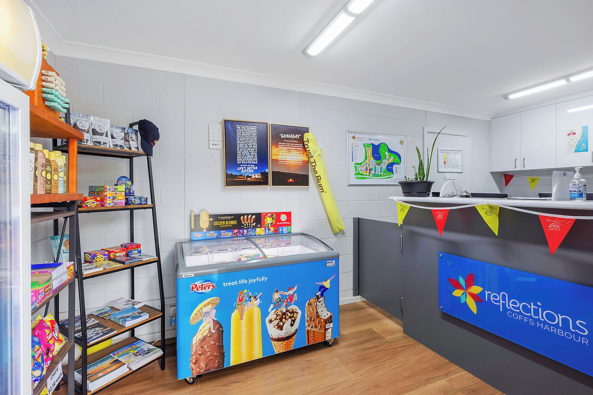 A modern ice cream parlor with a freezer display, shelves with snacks, and a counter with a sign for 'Reflections Coffs Harbour'.