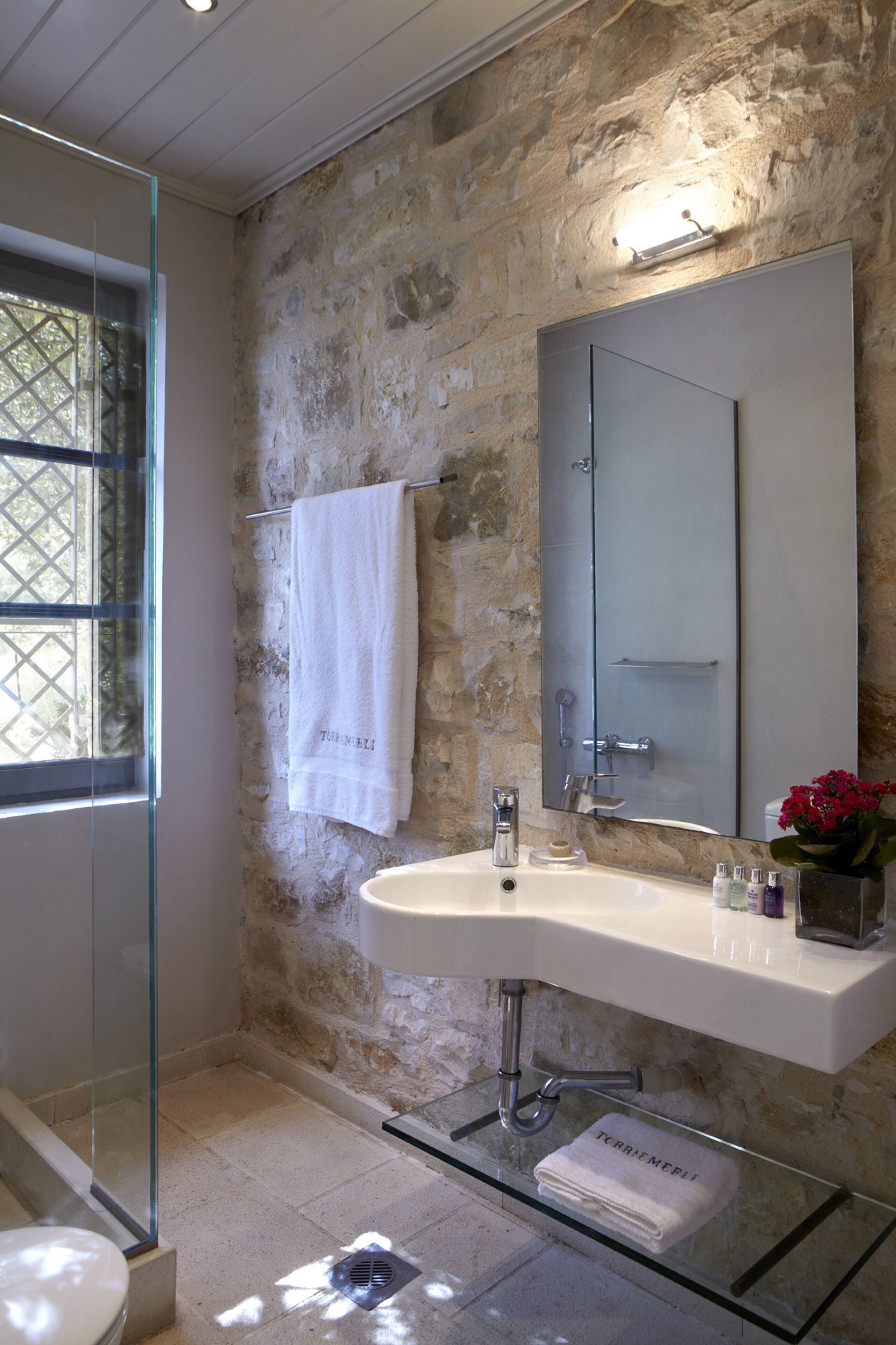A bathroom with a stone wall, a glass shower enclosure, a white sink, and a large mirror.