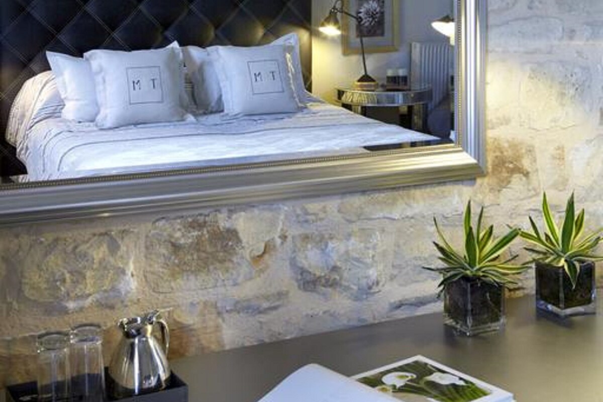 Une chambre avec un grand miroir, un lit avec une literie blanche, une table de chevet avec une lampe et deux plantes en pot.