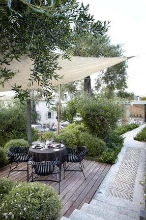 An outdoor dining area with a round table and chairs, surrounded by greenery and a wooden deck. - Torri E Merli Hotel by GHH (Paxos)