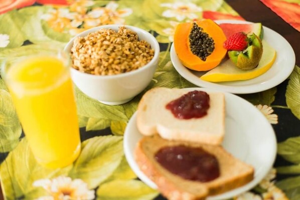 A breakfast table with a bowl of granola, a plate with a sliced papaya and kiwi, toast with jam, and a glass of orange juice. - Pousada Dom Fernandes (Cabo Frio)