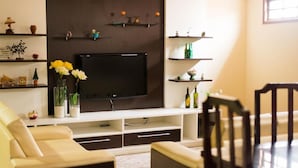A living room with a flat-screen TV mounted on a white entertainment unit, a beige sofa, a wooden dining table, and a dark wooden chair. - Pousada Dom Fernandes (Cabo Frio)