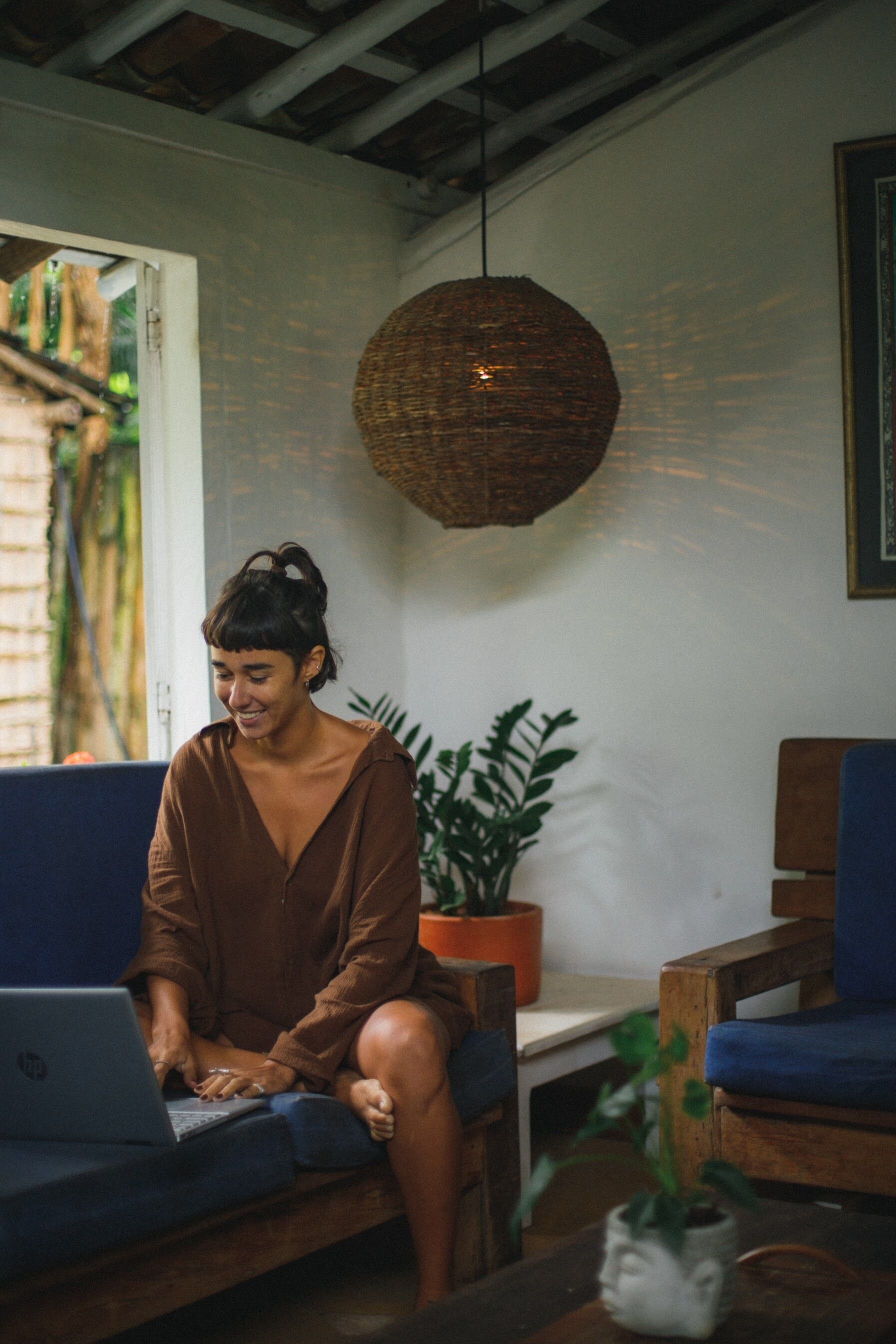 A person sitting on a blue couch using a laptop. There is a wicker hanging light fixture above and a potted plant to the right.