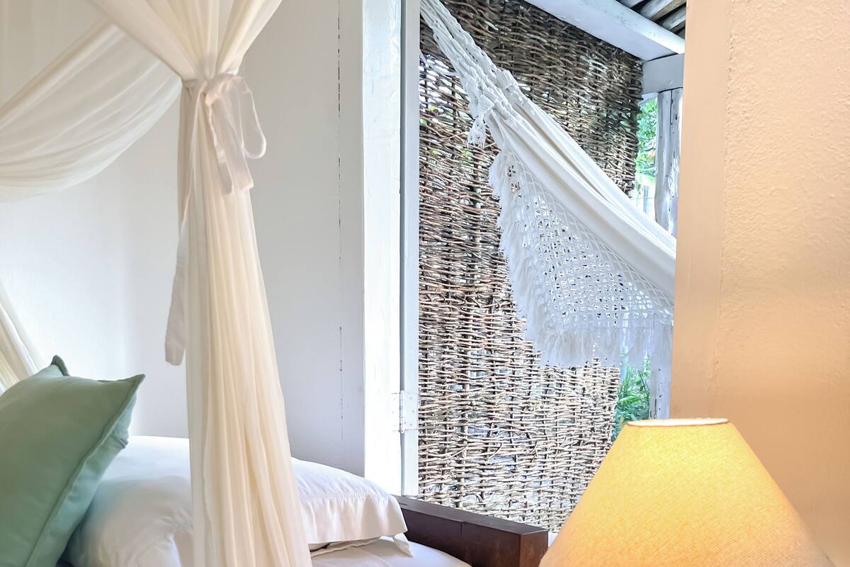Ein Schlafzimmer mit einem Bett, einem Nachttisch aus Holz, einer Lampe und Blick durch ein Fenster mit einem Gewebeschirm.