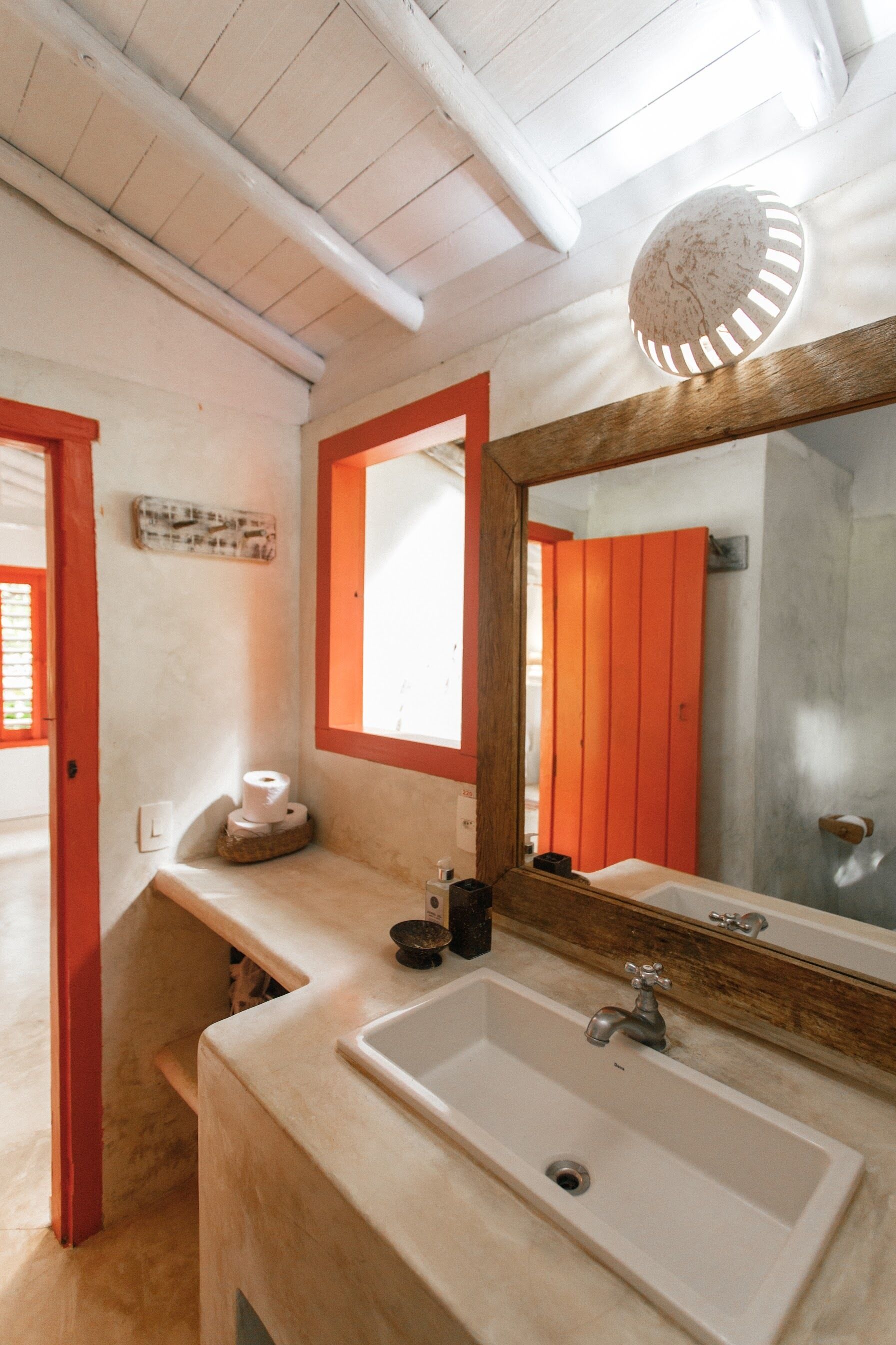 Un baño con un lavamanos blanco, un espejo con marco de madera y una ventana con marco rojo.