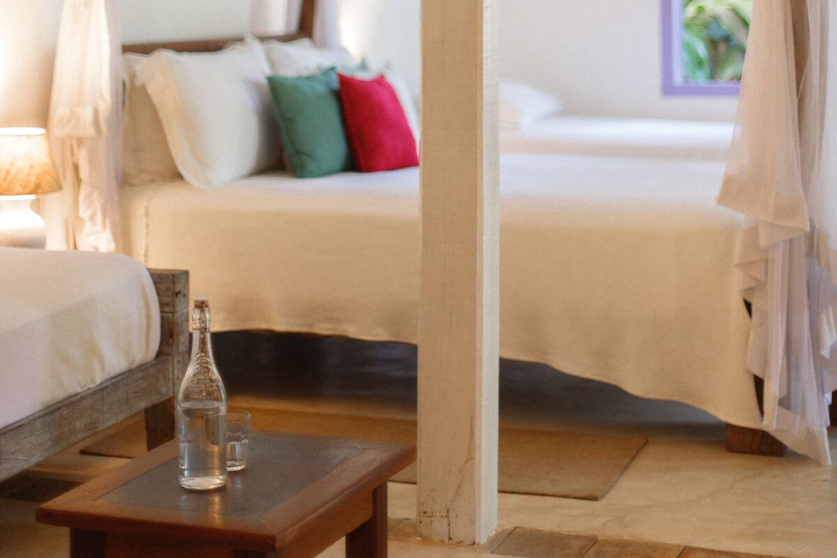 Ein Zimmer mit einem Himmelsbett, einem Nachttisch mit Flasche und Glas sowie einem Fenster mit Vorhängen.