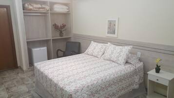 Um quarto com cama, uma cadeira, mesa de cabeceira, mesinha e um guarda-roupa, além de um quadro na parede.