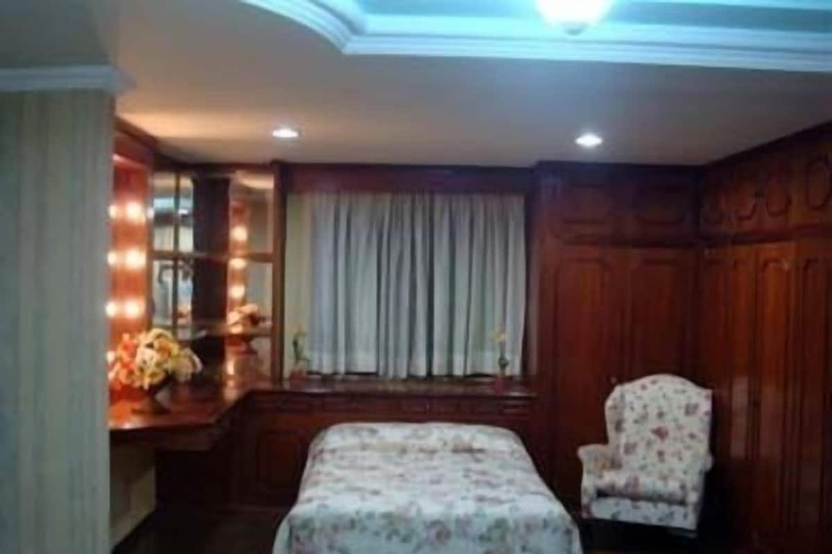 A bedroom with a bed, a chair, a vanity with a mirror, and a window with curtains.