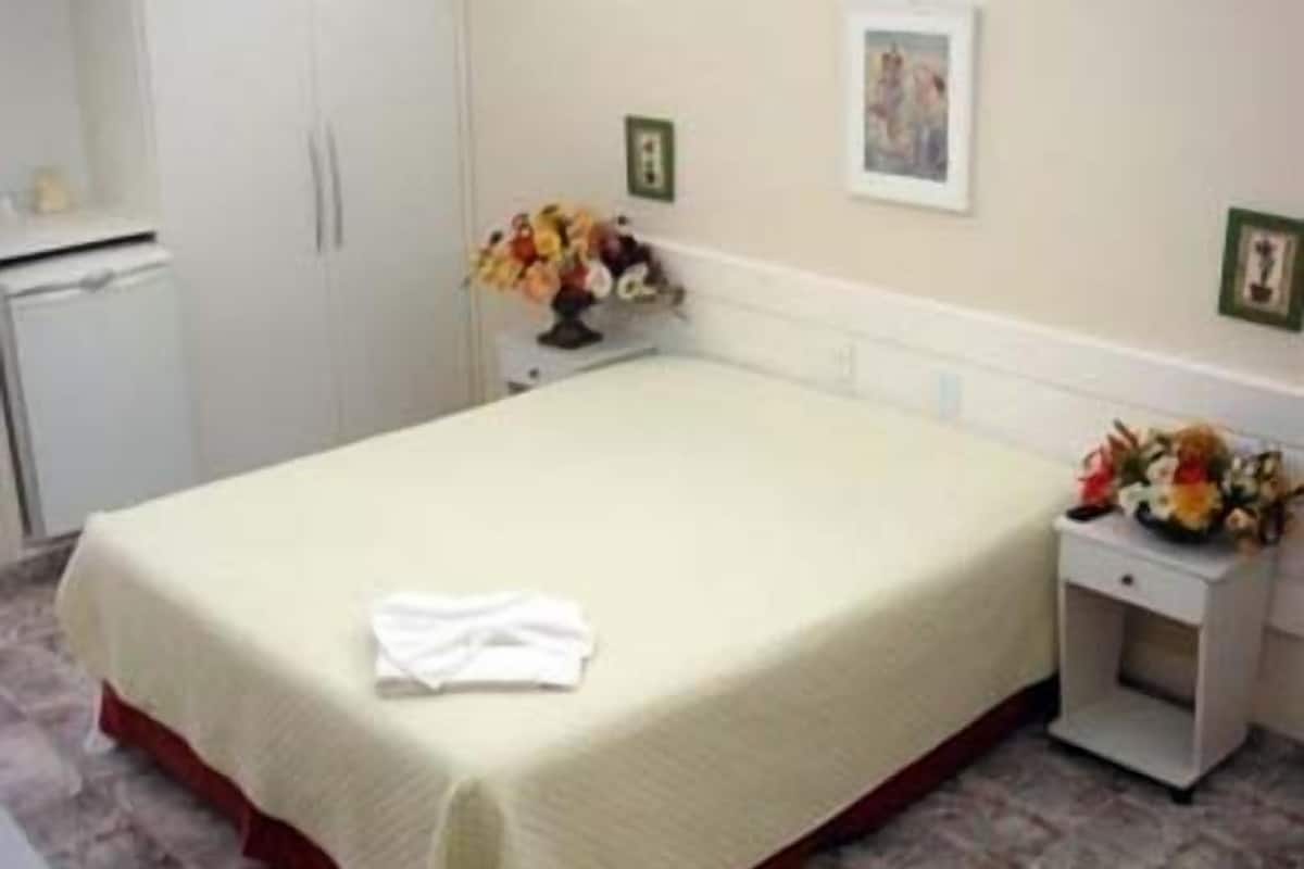 A single bed with a white bedspread and a red trim, a nightstand with a vase of flowers, and a small cabinet.