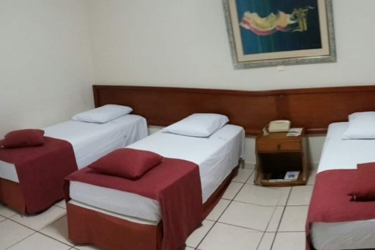 A hotel room with four single beds, each with a red cover and white pillow. There is a wooden headboard, a small wooden nightstand with a telephone, and a framed picture on the wall.