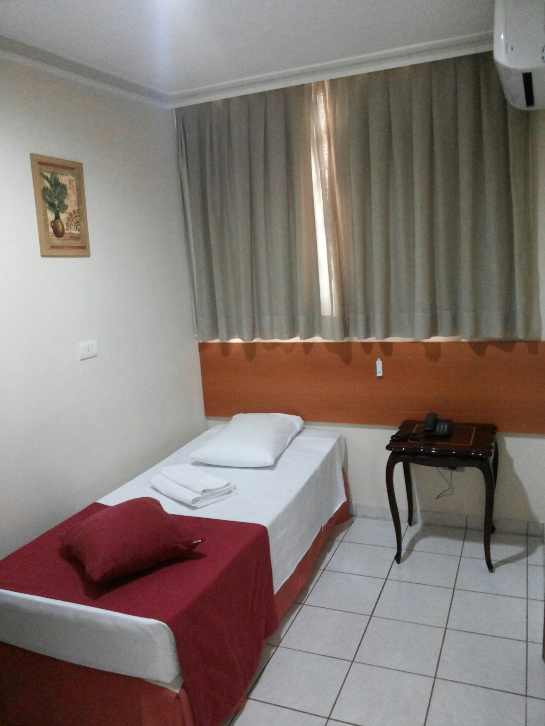 A single bed with a red blanket, a bedside table with a phone, and a window with curtains.