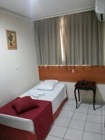 Una cama individual con una manta roja, una mesita de noche con un teléfono y una ventana con cortinas.