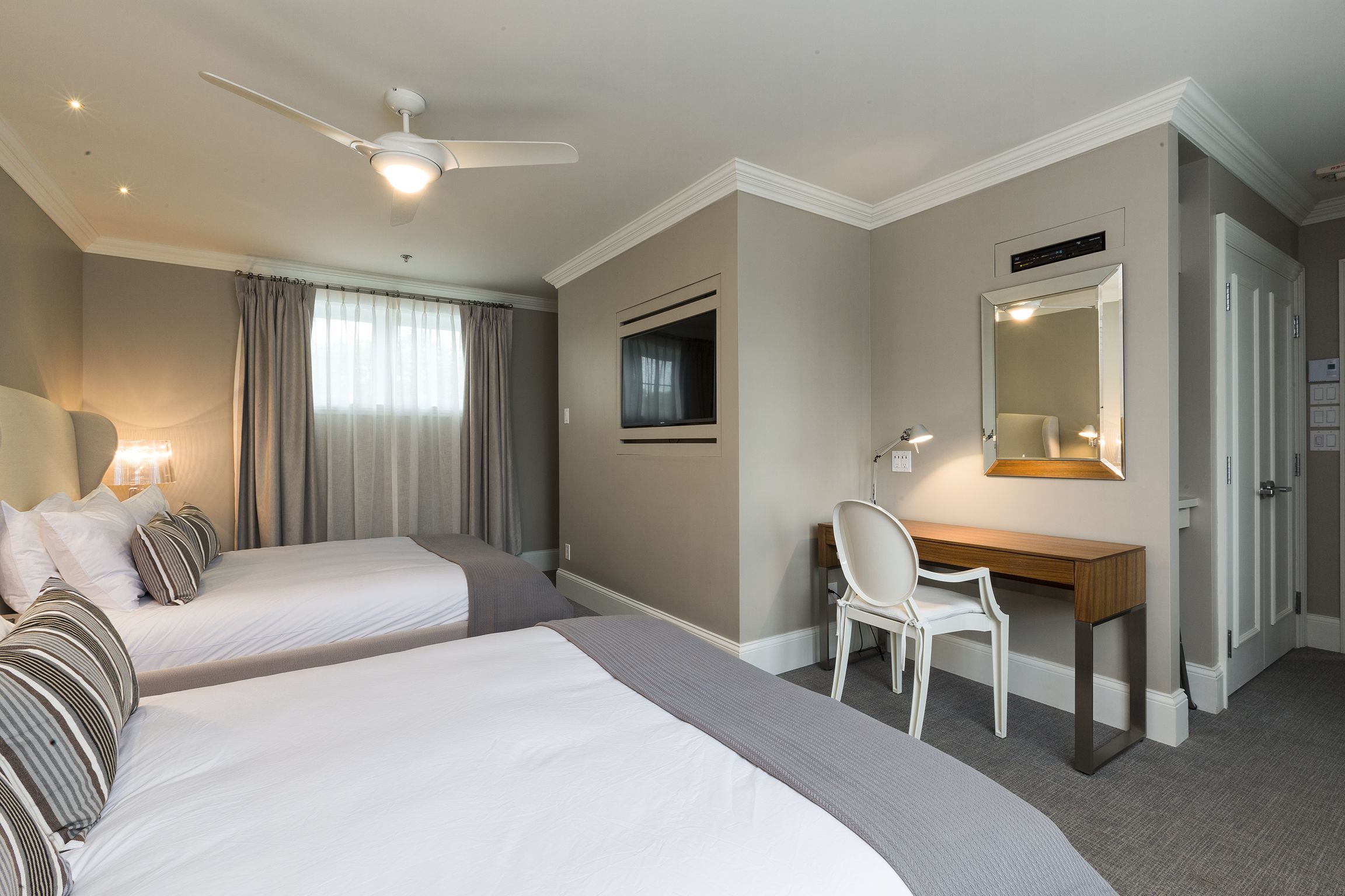 Habitación de hotel con dos camas, un escritorio, una silla, ventilador de techo, televisión y espejo.