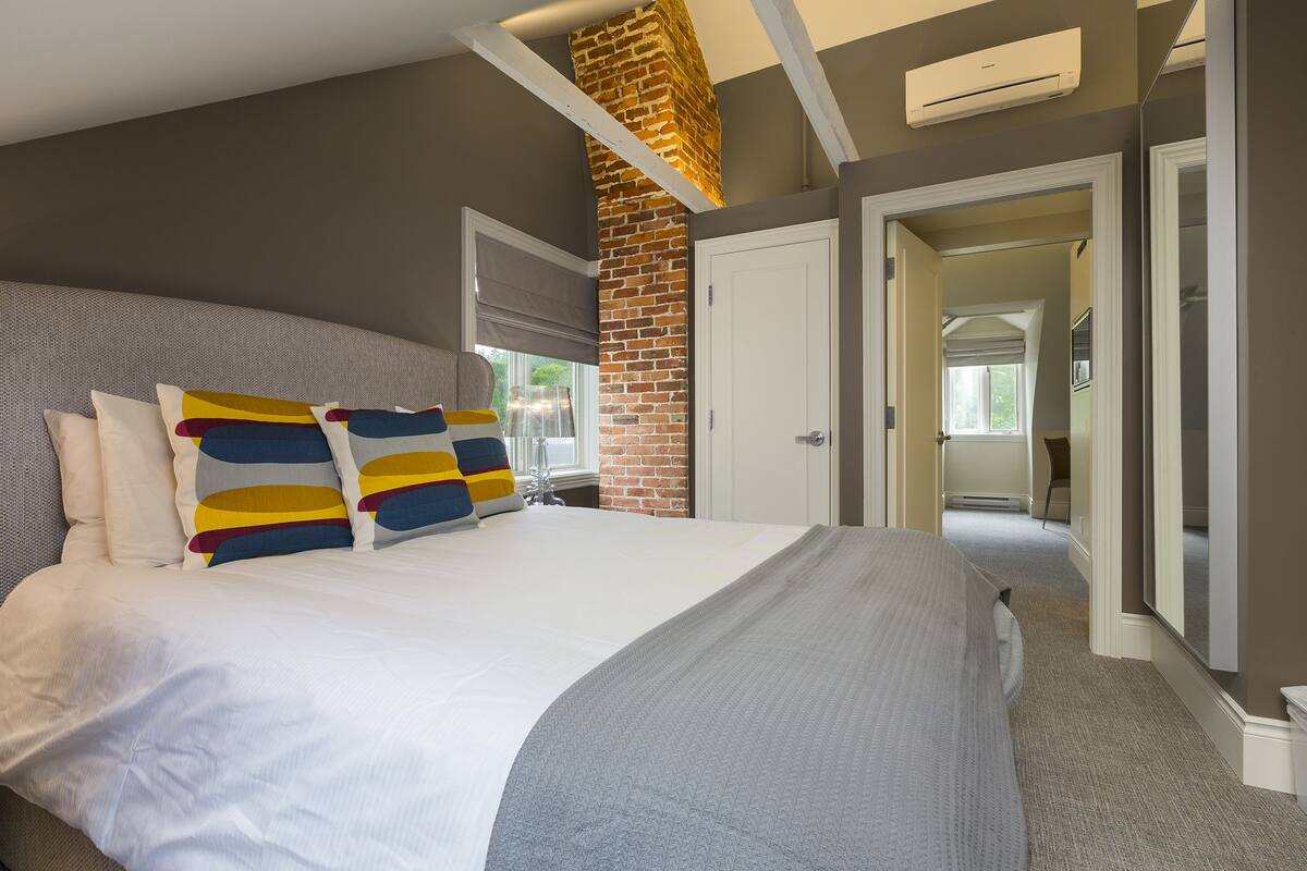 A modern bedroom with a large bed, a brick accent wall, and a hallway leading to another room.