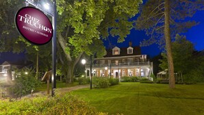 A well-lit bistro with a large marquee sign, surrounded by lush greenery and a large house in the background. - Chez Truchon (La Malbaie)