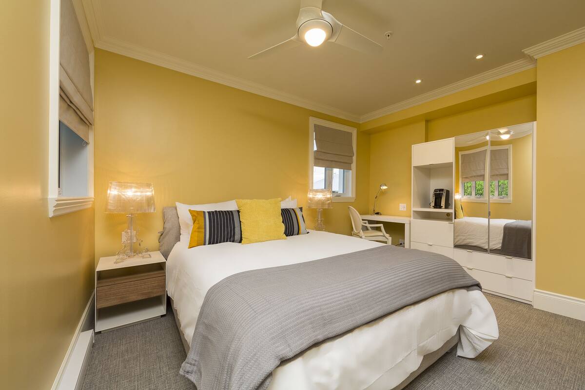 A hotel room with a bed, bedside table, lamp, mirror, and a desk with a chair.