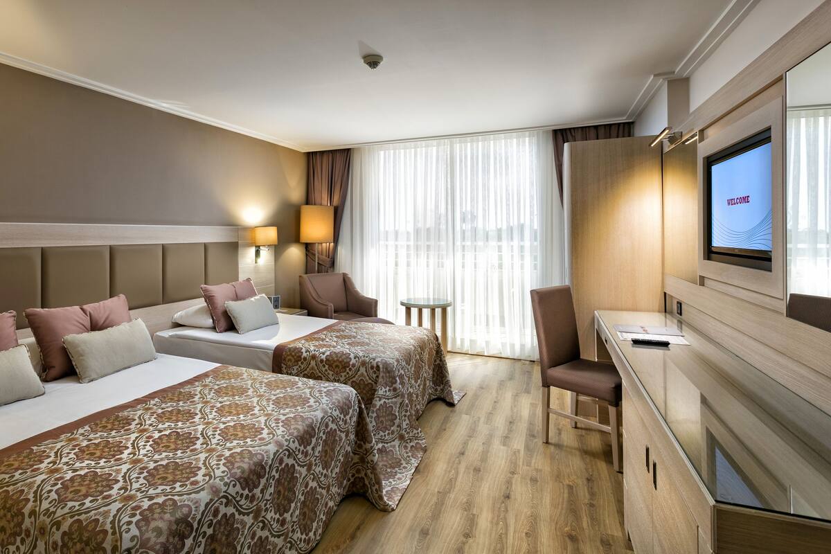 Une chambre d’hôtel avec deux lits, un bureau, une chaise et un téléviseur affichant le mot « BIENVENUE ».