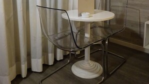 A small, clear glass table with a white base, surrounded by clear glass chairs, against a backdrop of white curtains and a patterned wall. - 7MOTEL - SETTIMOTEL (Settimo Milanese)