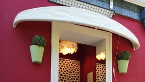 A red building entrance with a white awning, two potted plants, a small table with a plant, and a decorative wall. - 7MOTEL - SETTIMOTEL (Settimo Milanese)