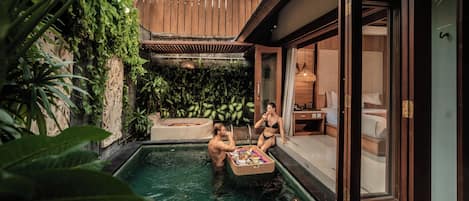 Un área de piscina privada con terraza de madera, abundante vegetación y una habitación con paredes de vidrio y una cama.