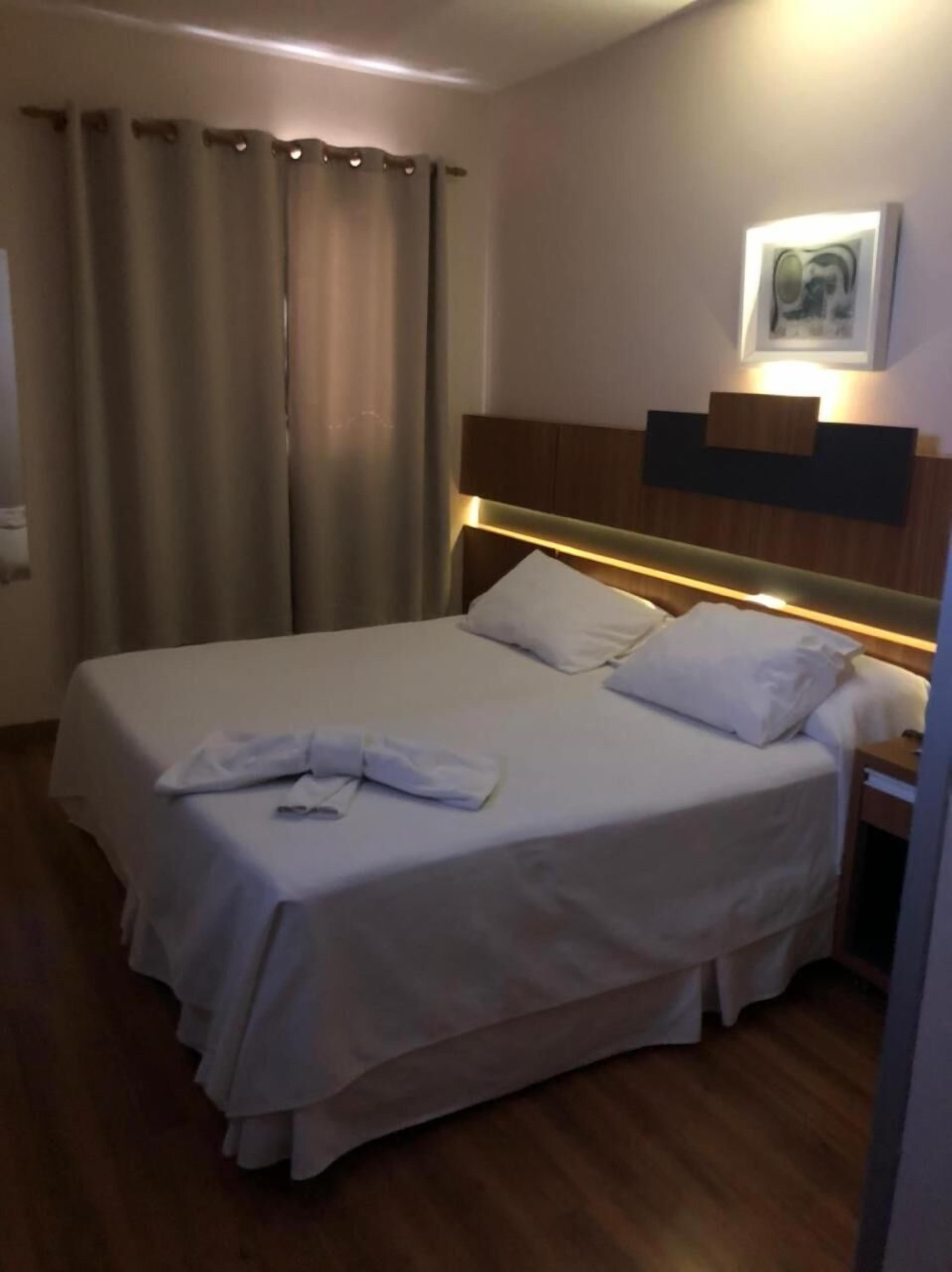 Ein Hotelzimmer mit einem Bett, Vorhängen, einem Kopfteil, einer Wandleuchte und einem gerahmten Bild.