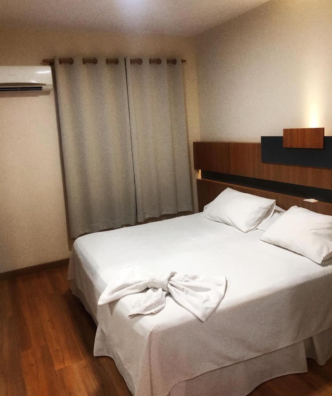 Una cama bien hecha con sábanas blancas y una toalla doblada, cabecera de madera y unidad de aire acondicionado en la pared.
