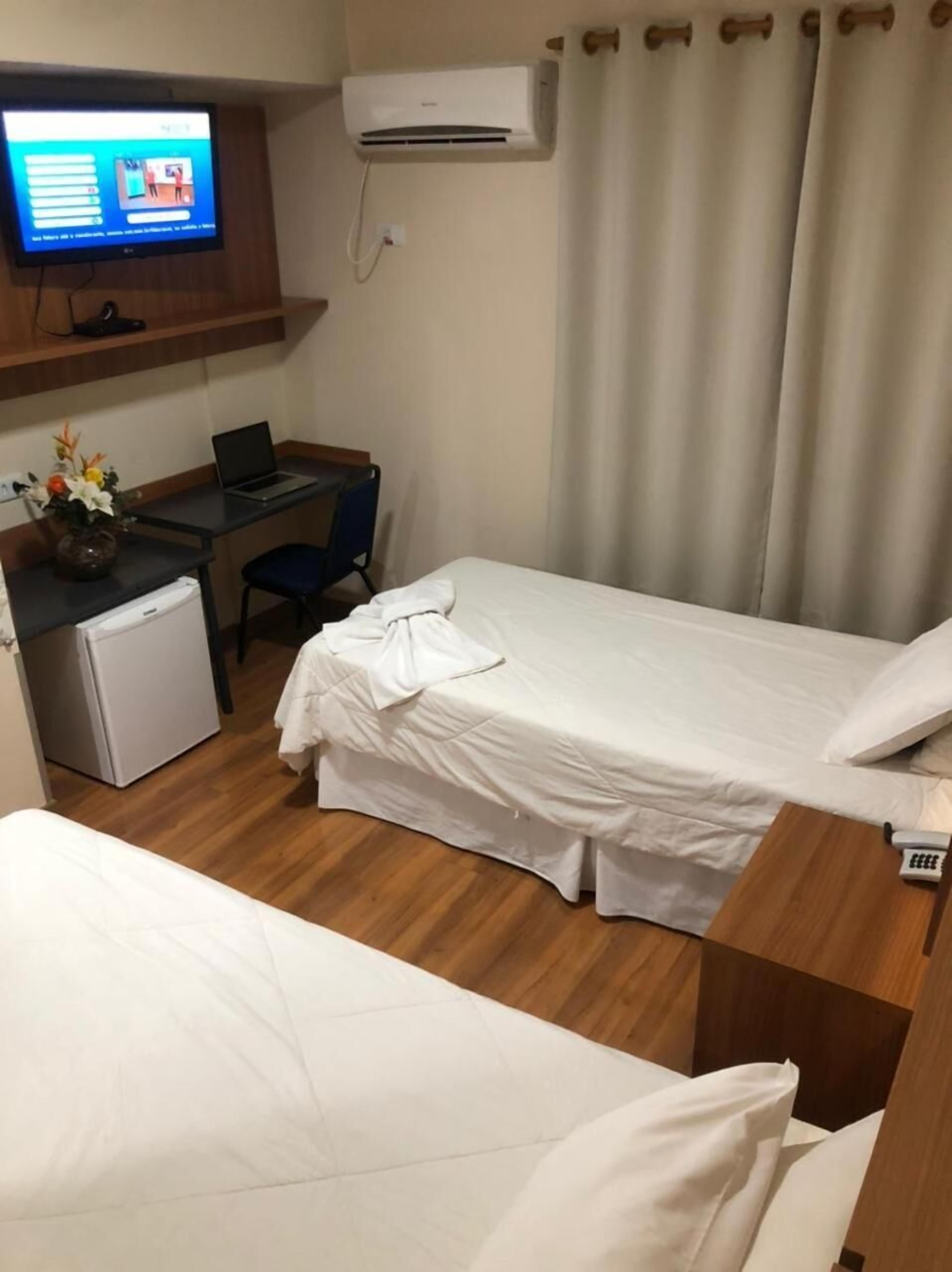 Een hotelkamer met een bed, een bureau met een laptop, een koelkast, een televisie aan de muur en een raam met gordijnen.