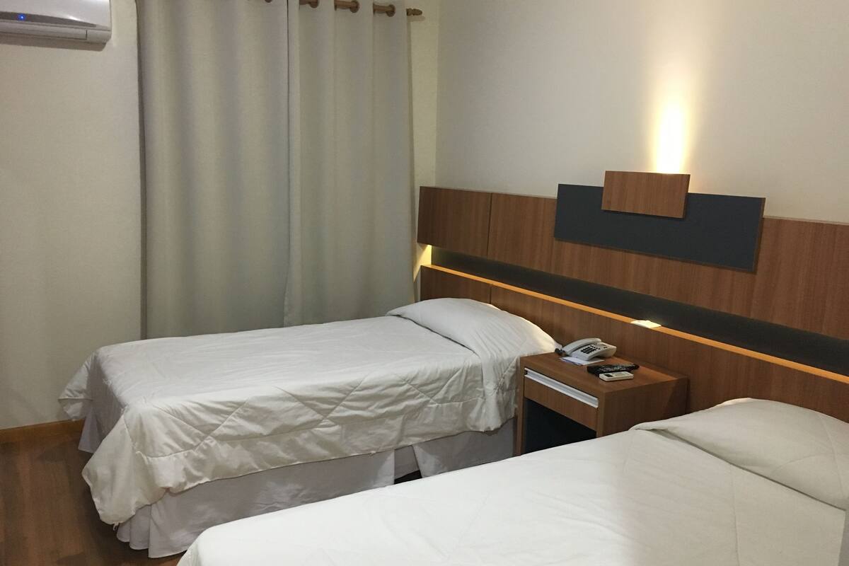 Une chambre d’hôtel avec deux lits simples, une table de chevet avec un téléphone et une télécommande, et un luminaire fixé au mur.