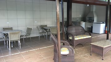 Un patio exterior con mobiliario de mimbre, piso de baldosas y una zona de cocina al fondo.