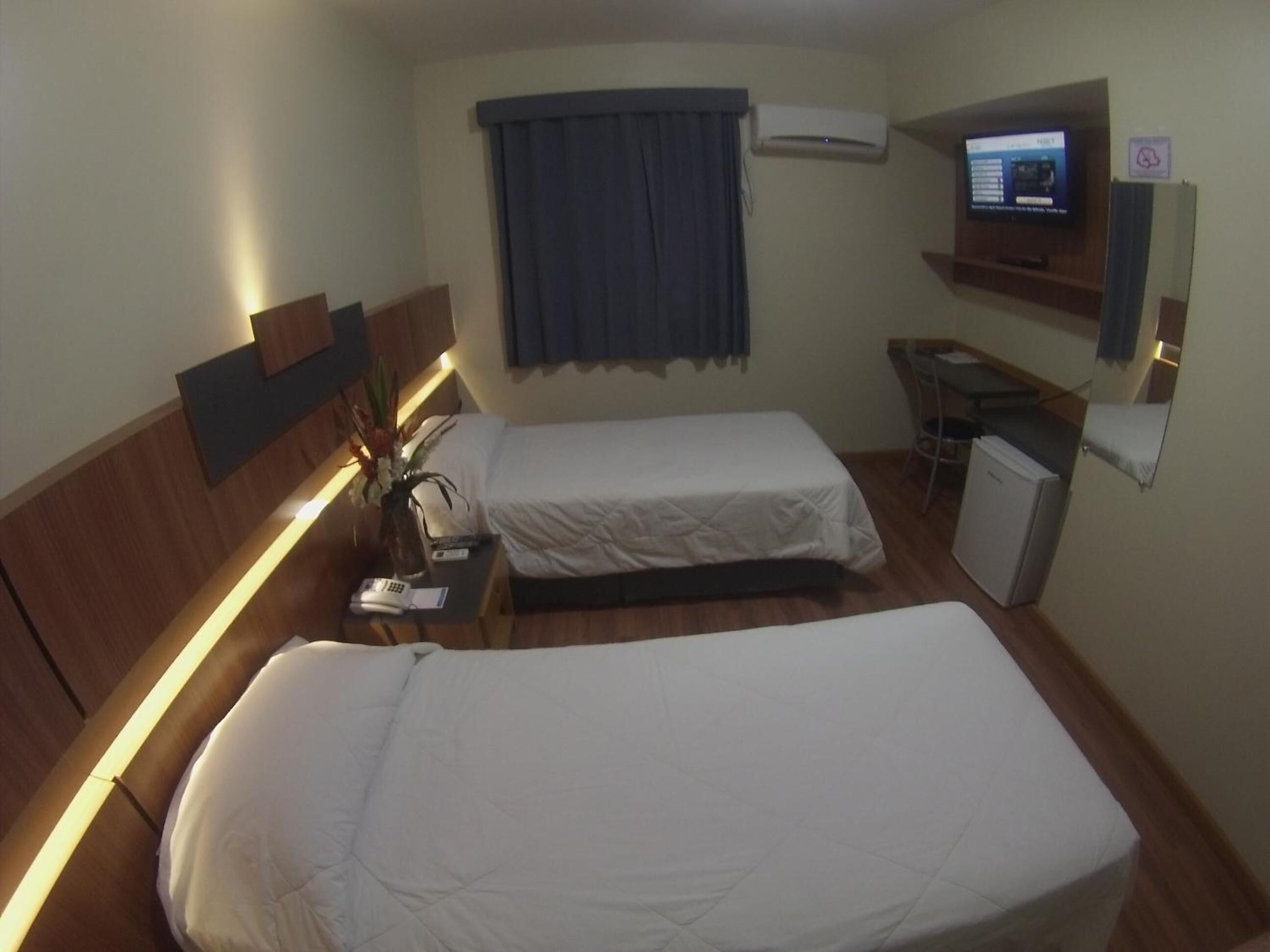 A hotel room with two beds, a desk, a small refrigerator, an air conditioning unit, and a television on the wall.