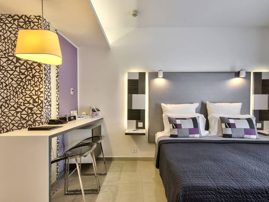 Ett modernt hotellrum med ett skrivbord, stolar, en säng med ett mörkt täcke och tapet med geometriskt mönster.