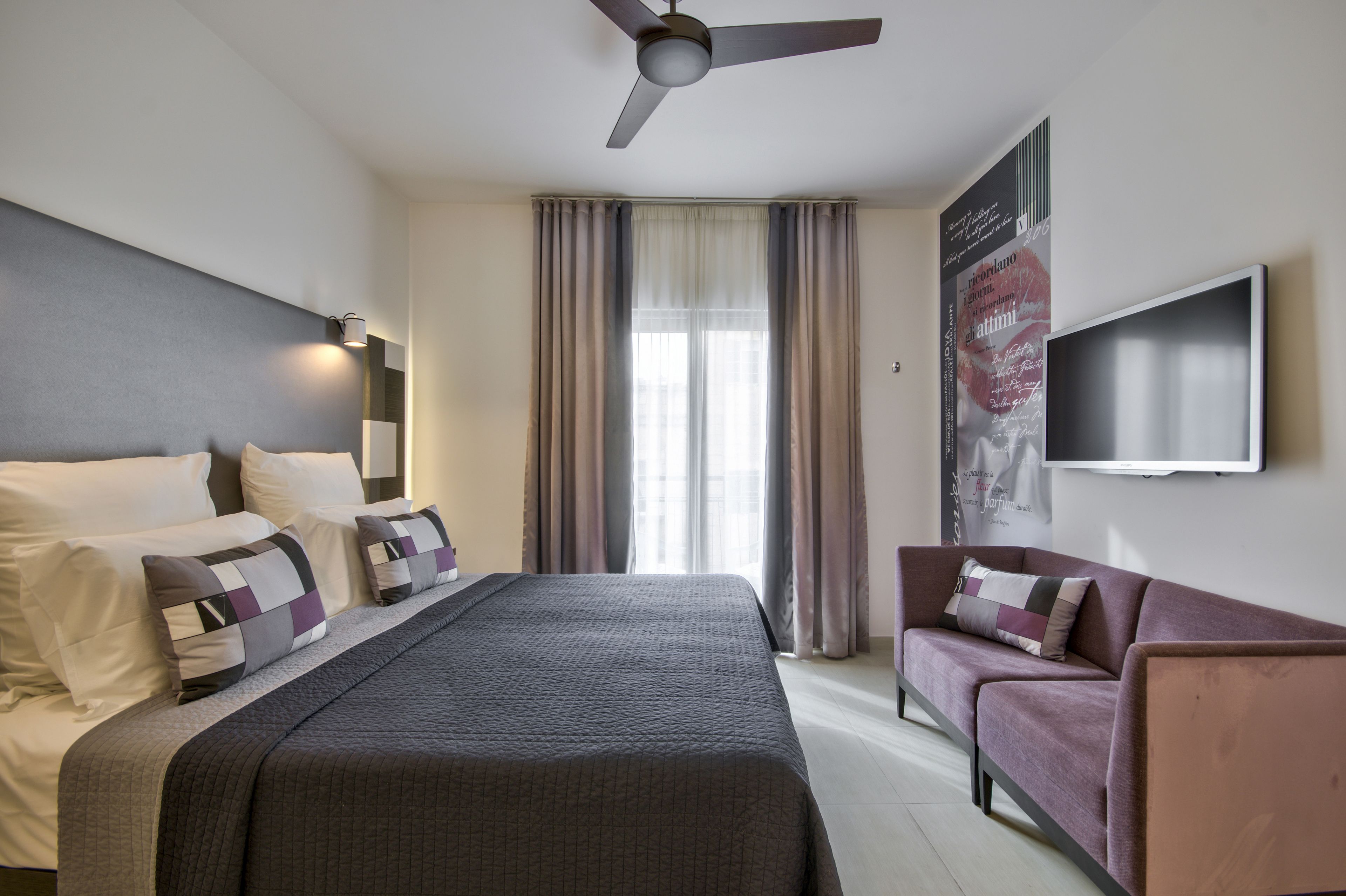 A hotel room with a bed, a sofa, a ceiling fan, a TV, and a window with curtains.