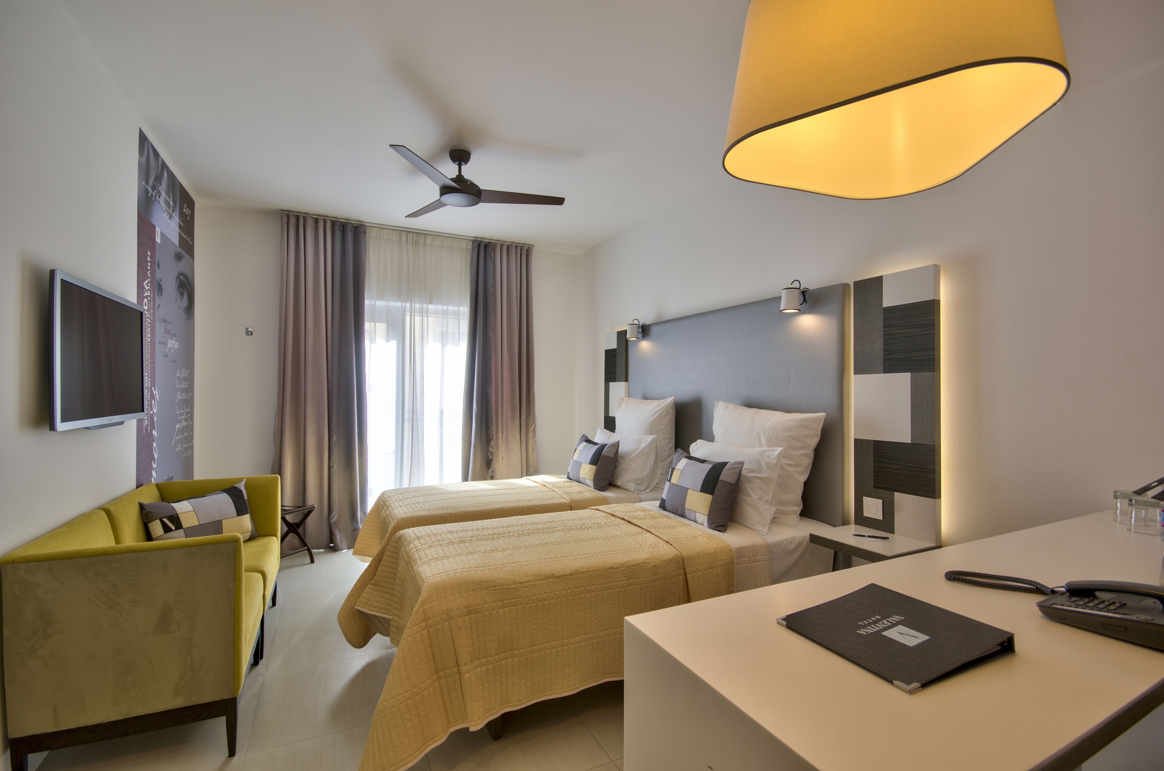 A hotel room with two beds, a desk, a chair, a TV, and a ceiling fan.