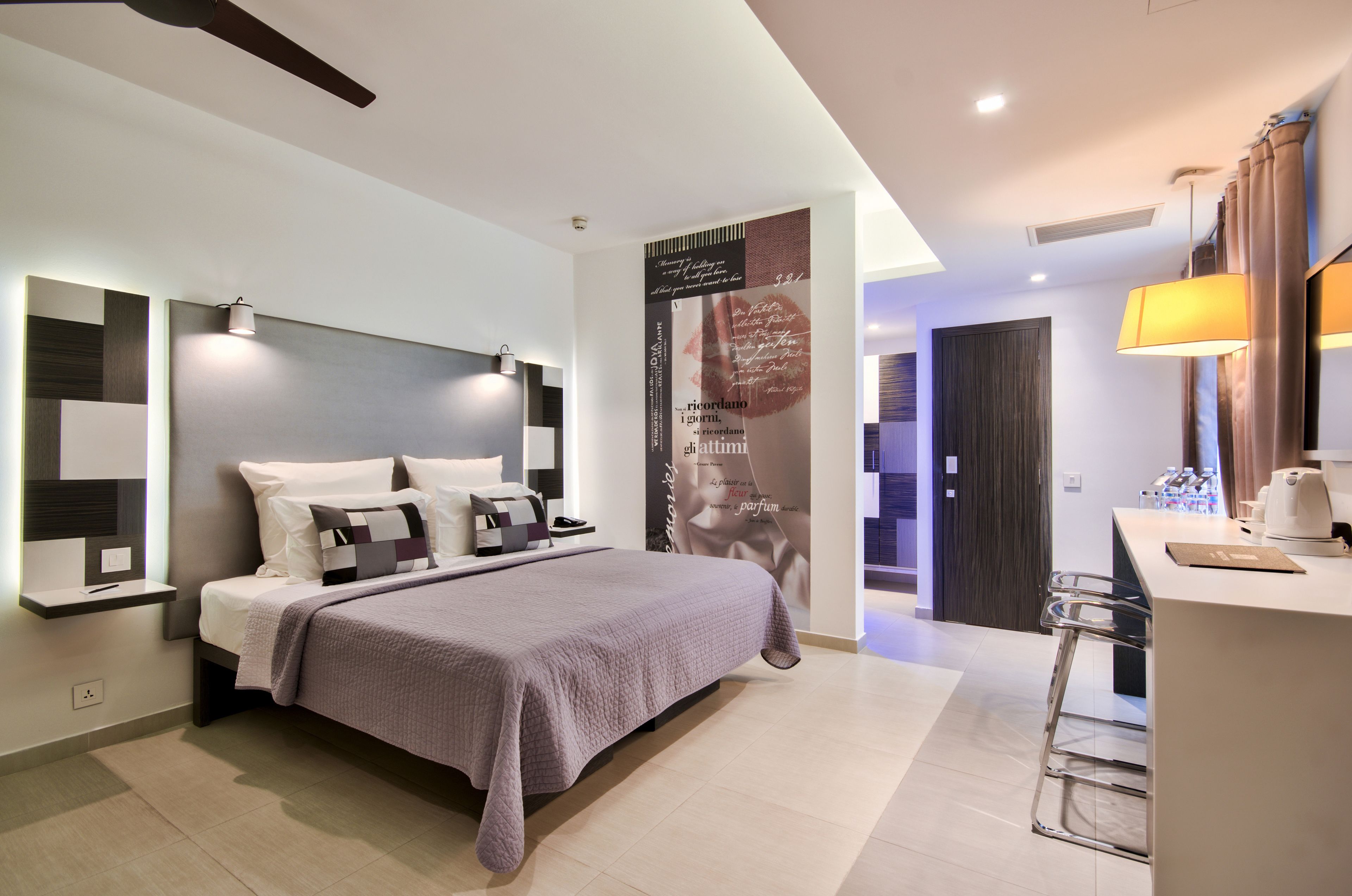 Un dormitorio moderno con cama, mesitas de noche, escritorio con silla, y baño visible a través de una puerta abierta.