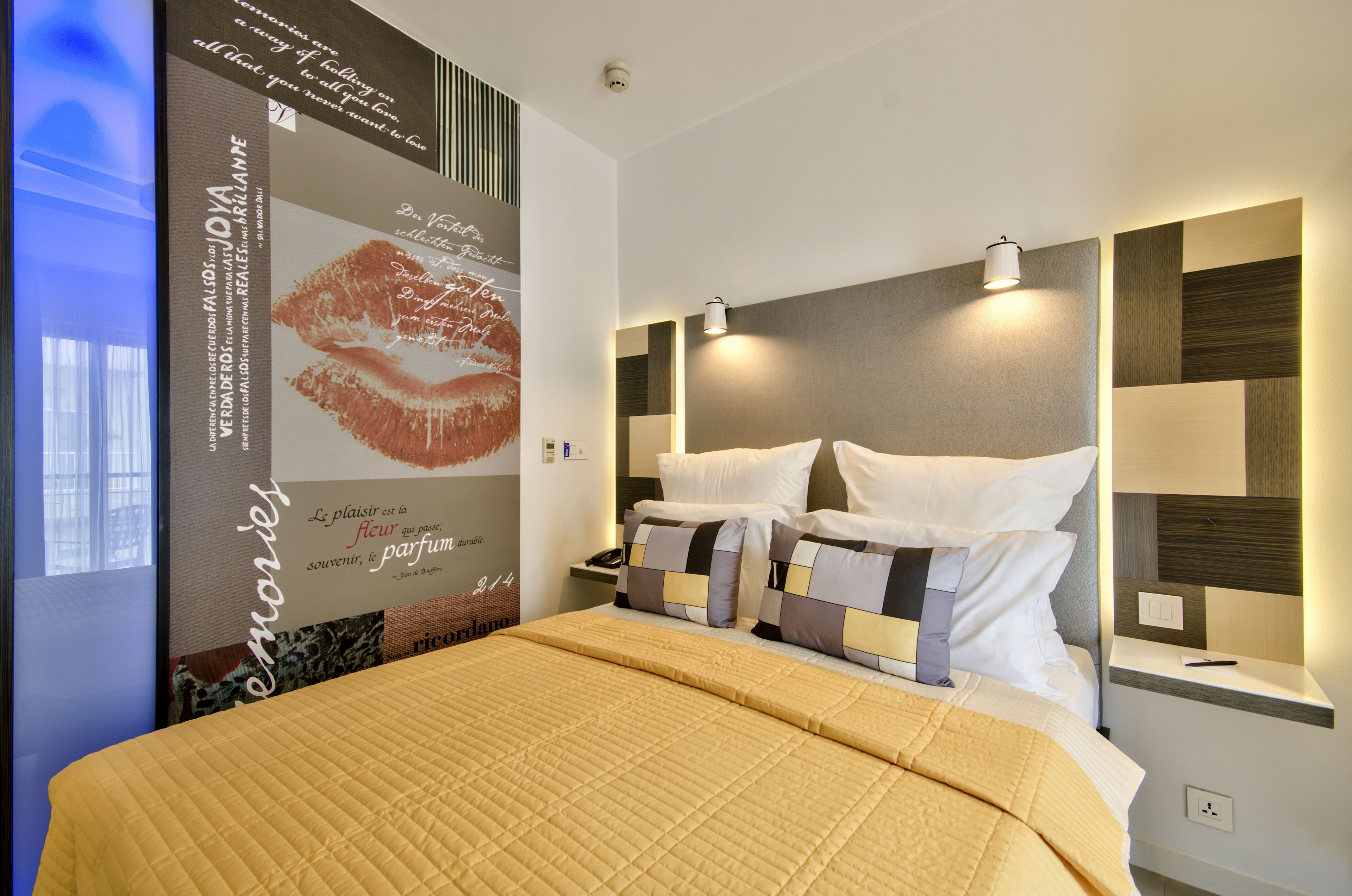 A modern hotel room with a large bed, a nightstand, a wall-mounted lamp, and a decorative wall with text and a lipstick print.