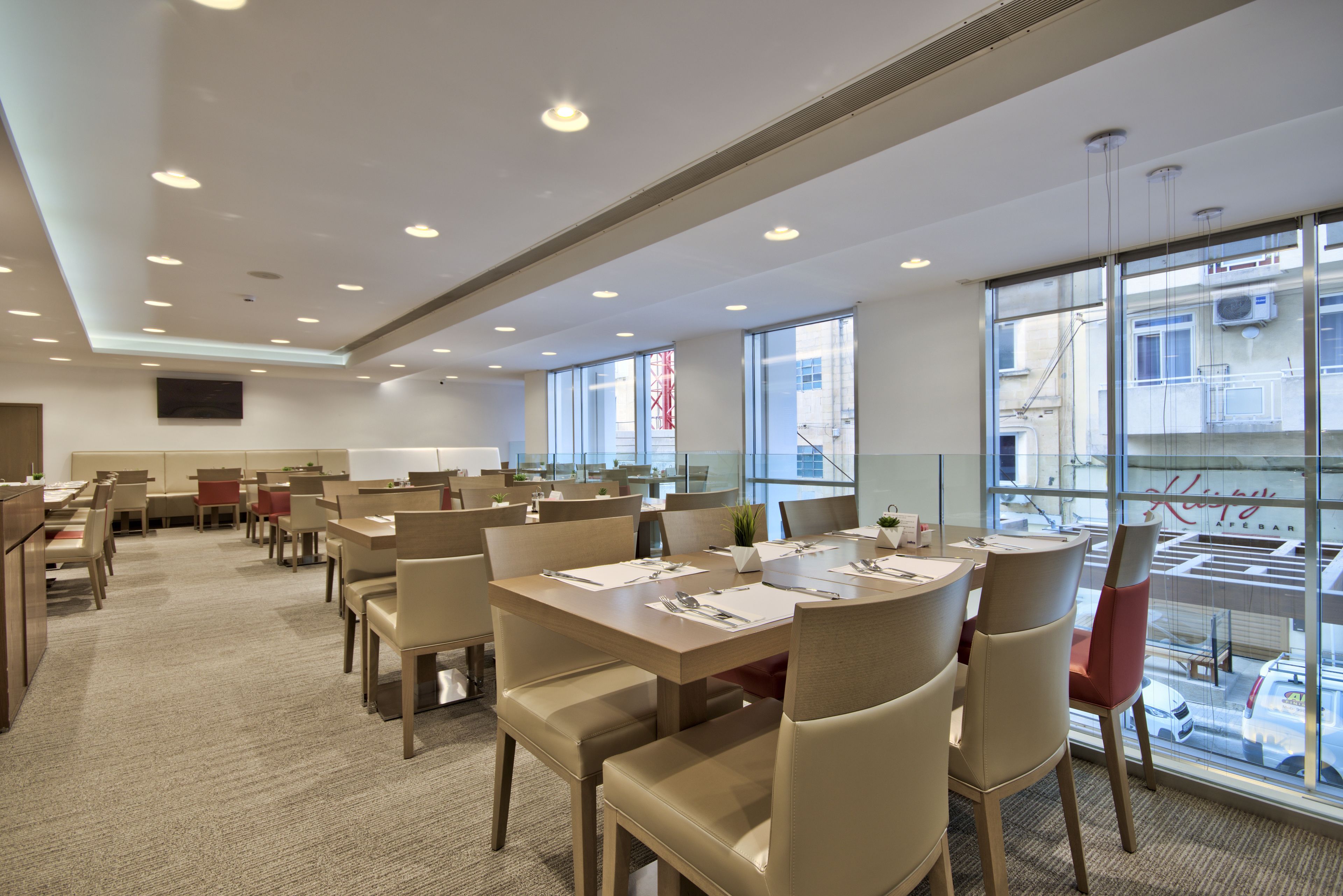 A modern dining area with multiple tables and chairs, large windows with a view of the city, and a flat-screen TV mounted on the wall.