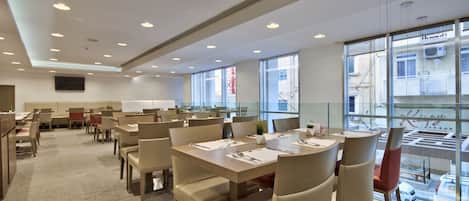 A modern dining area with multiple tables and chairs, large windows with a view of the city, and a flat-screen TV mounted on the wall.