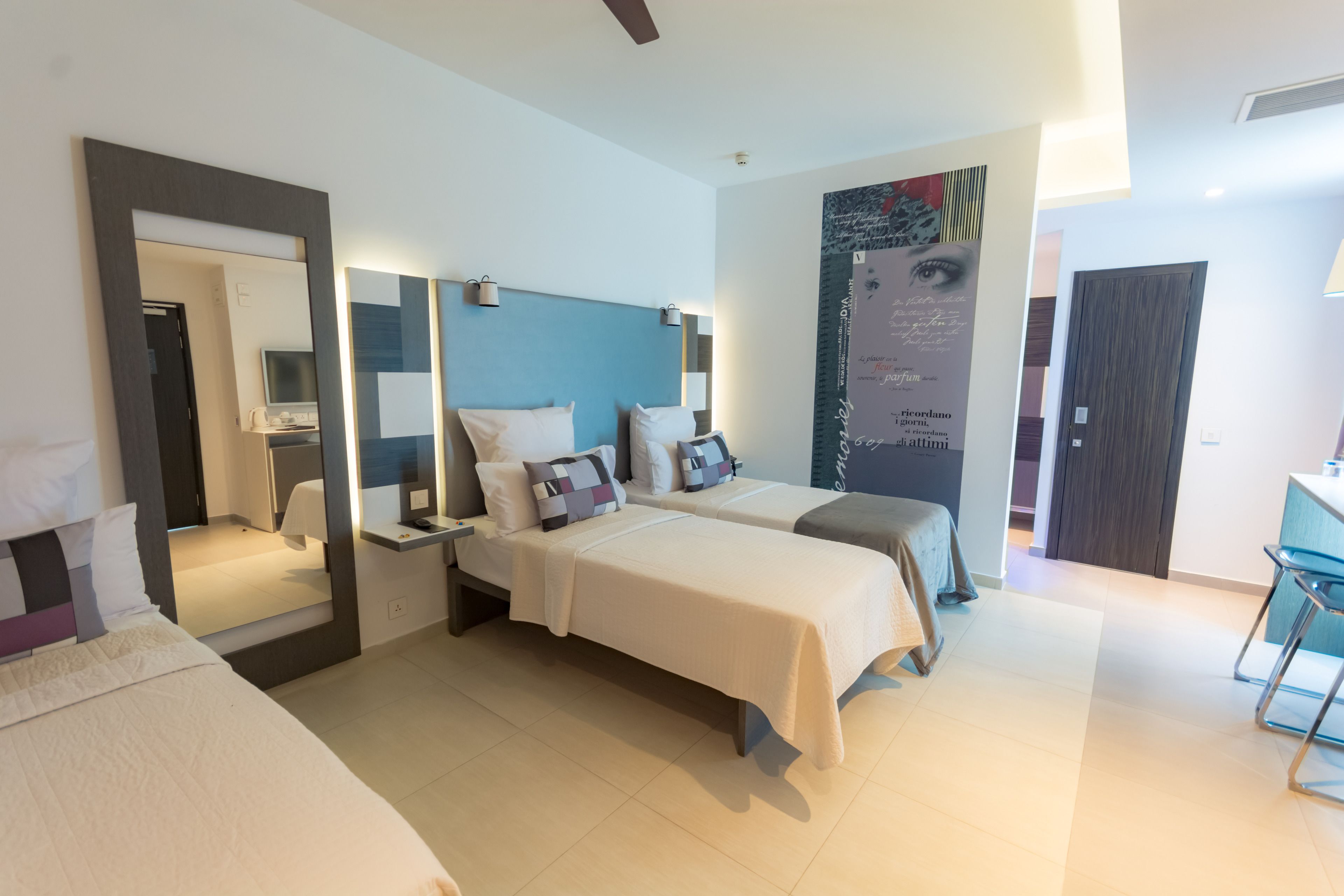 A modern hotel room with two beds, a large mirror, a wall-mounted TV, and a bathroom visible through an open door.