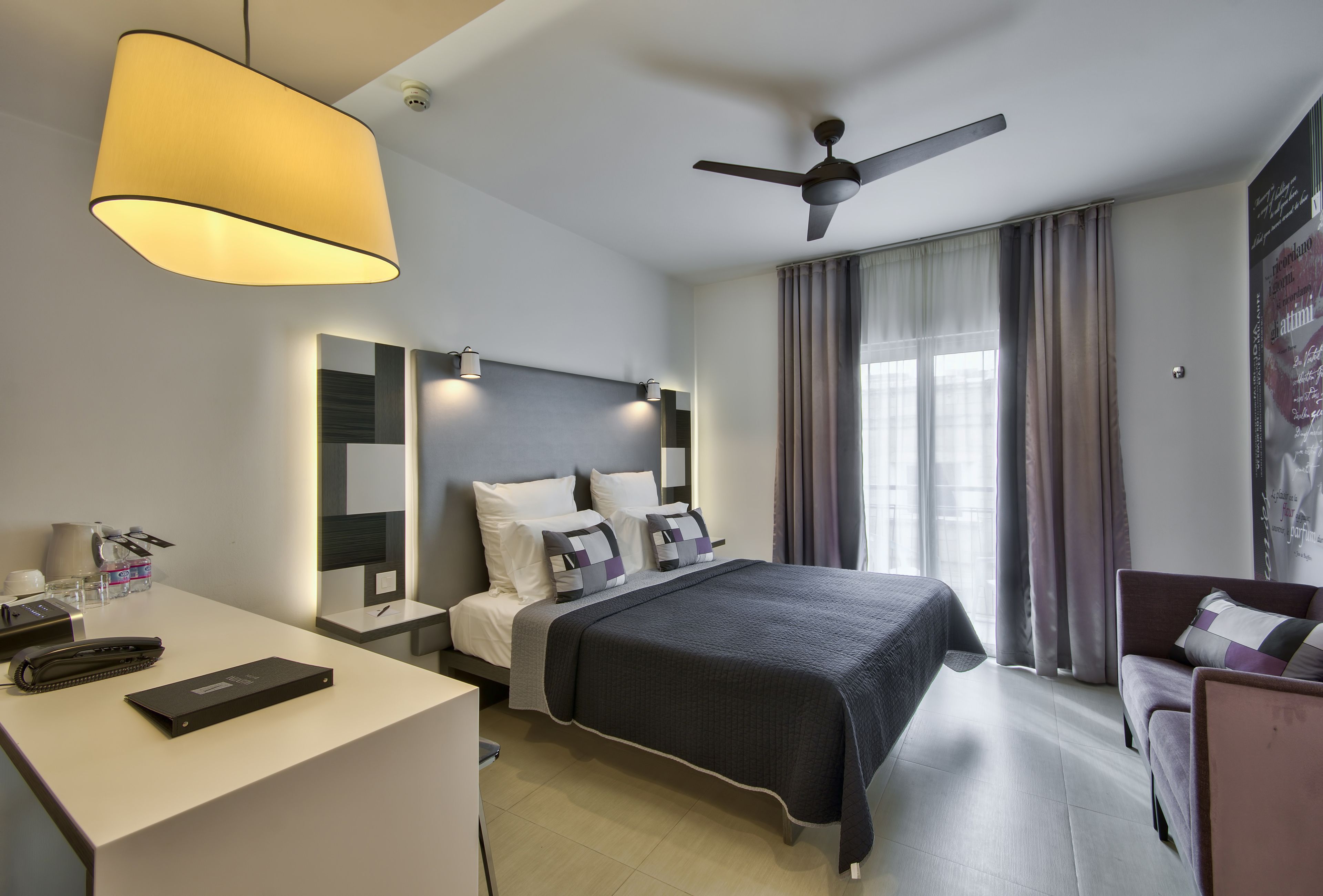 A modern hotel room with a bed, desk, and chair, featuring a ceiling fan, large windows with curtains, and a light fixture.
