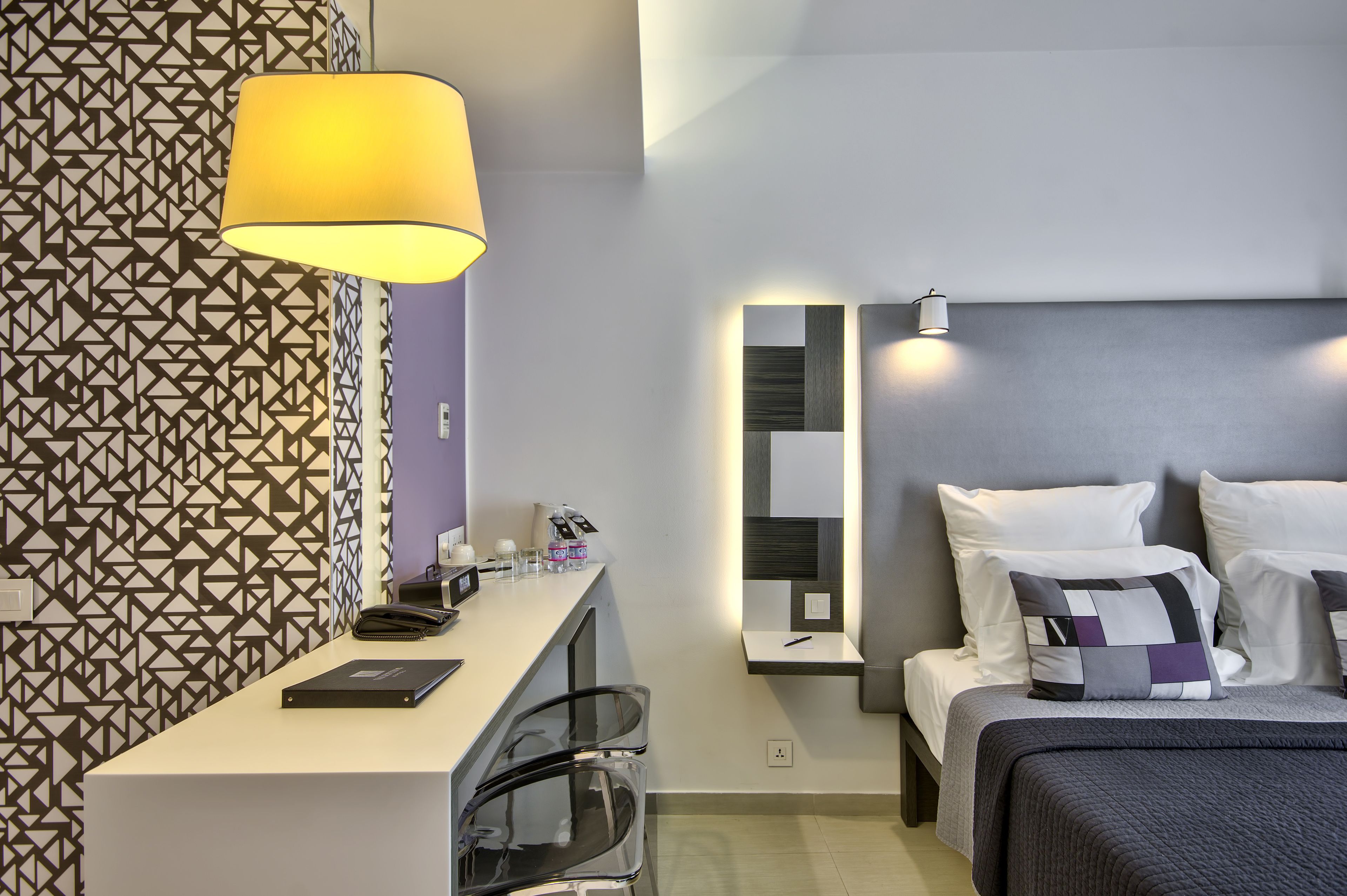 A modern hotel room with a desk, chairs, a bed with multiple pillows, a patterned wall, and a large lamp.