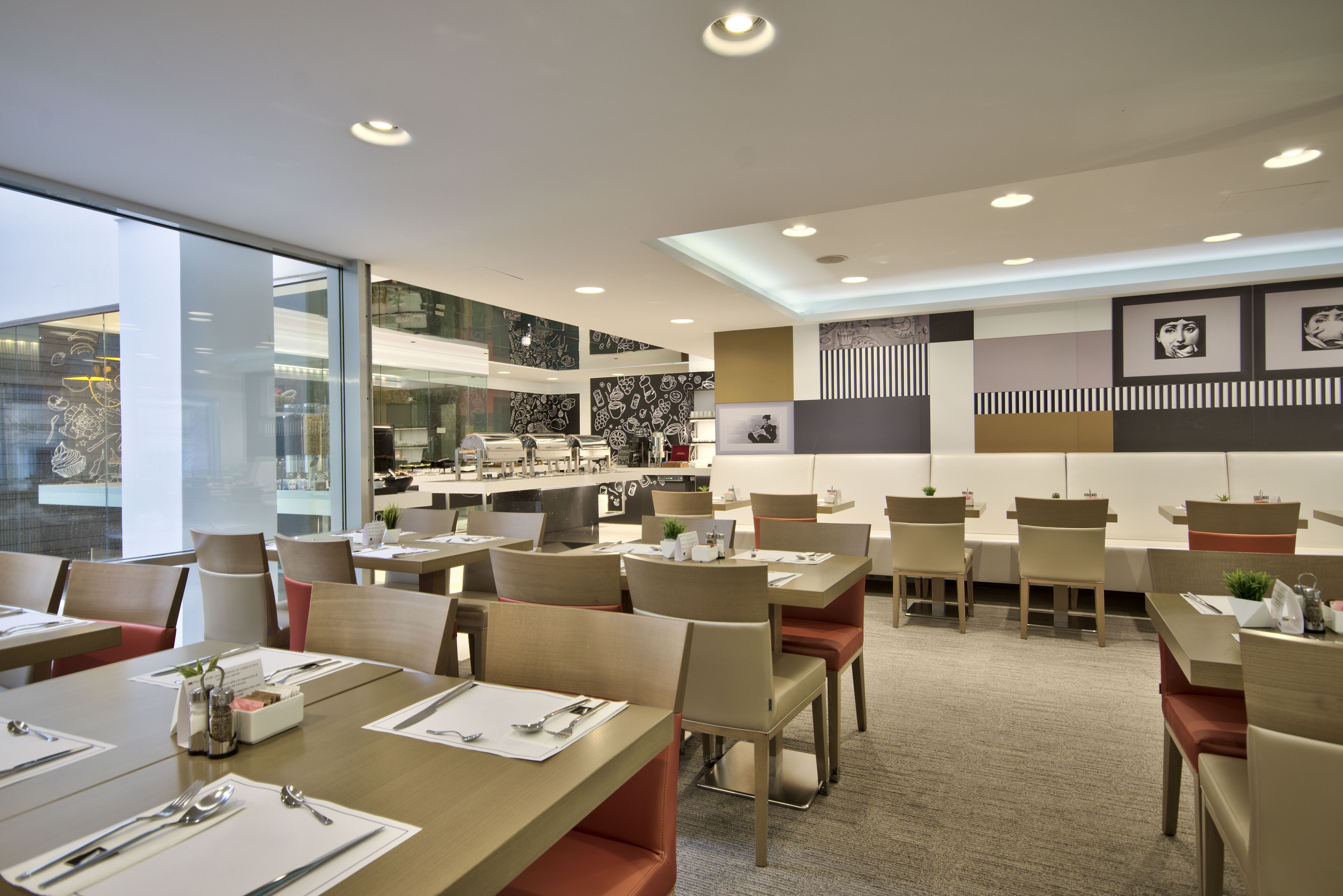 A modern restaurant with multiple dining tables, chairs, and a kitchen area visible through glass partitions.