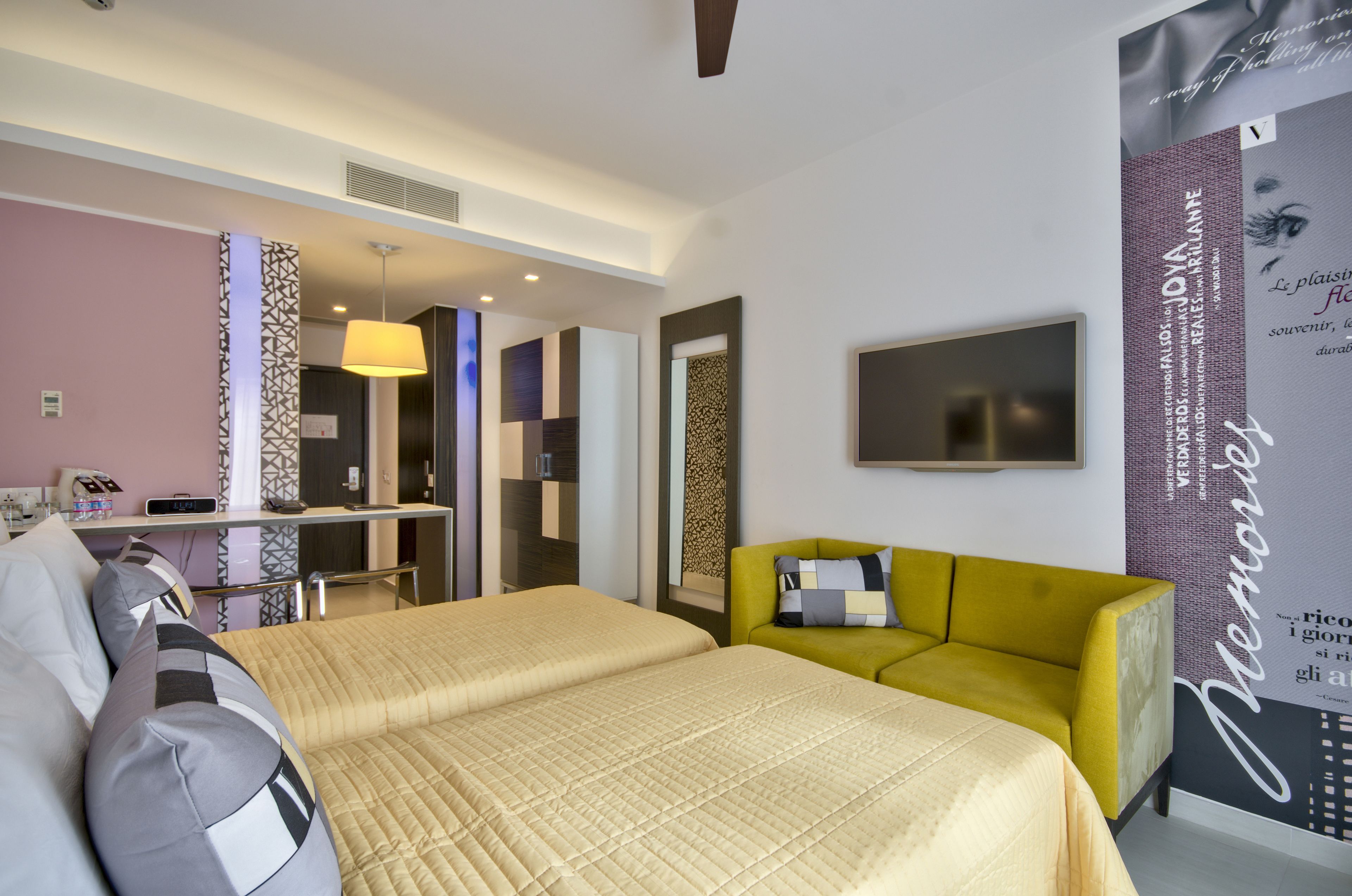 Une chambre d’hôtel avec un lit, un fauteuil jaune, un bureau avec une lampe et un téléviseur fixé au mur.