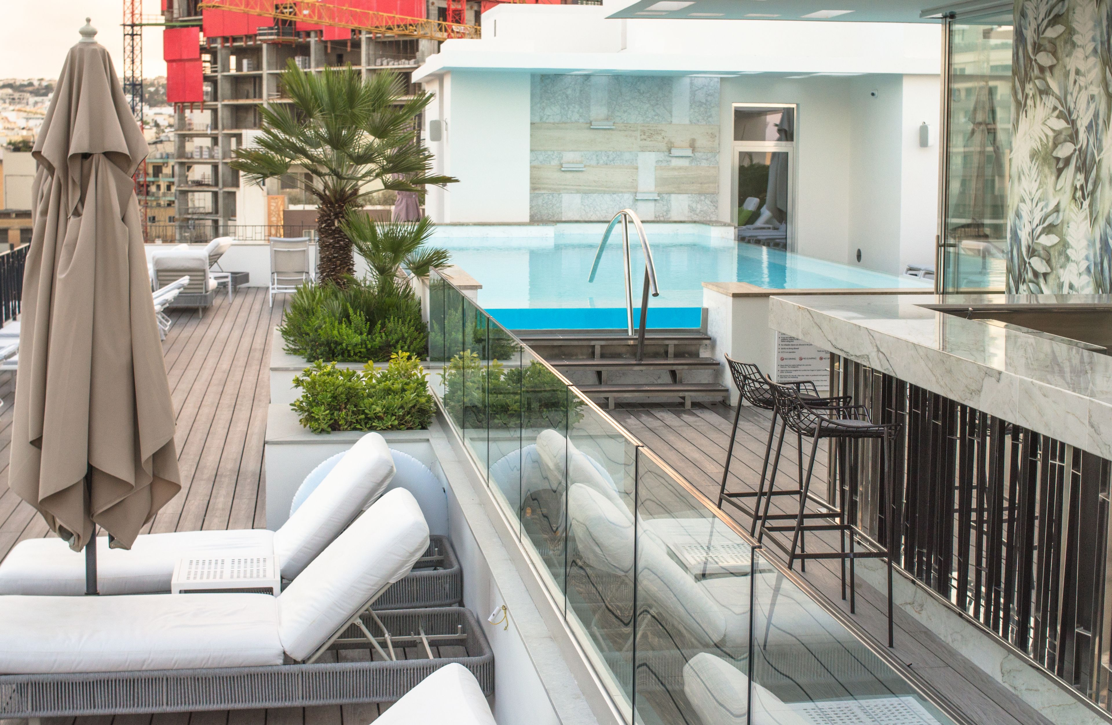 A rooftop pool area with lounge chairs, a bar, and a glass railing.