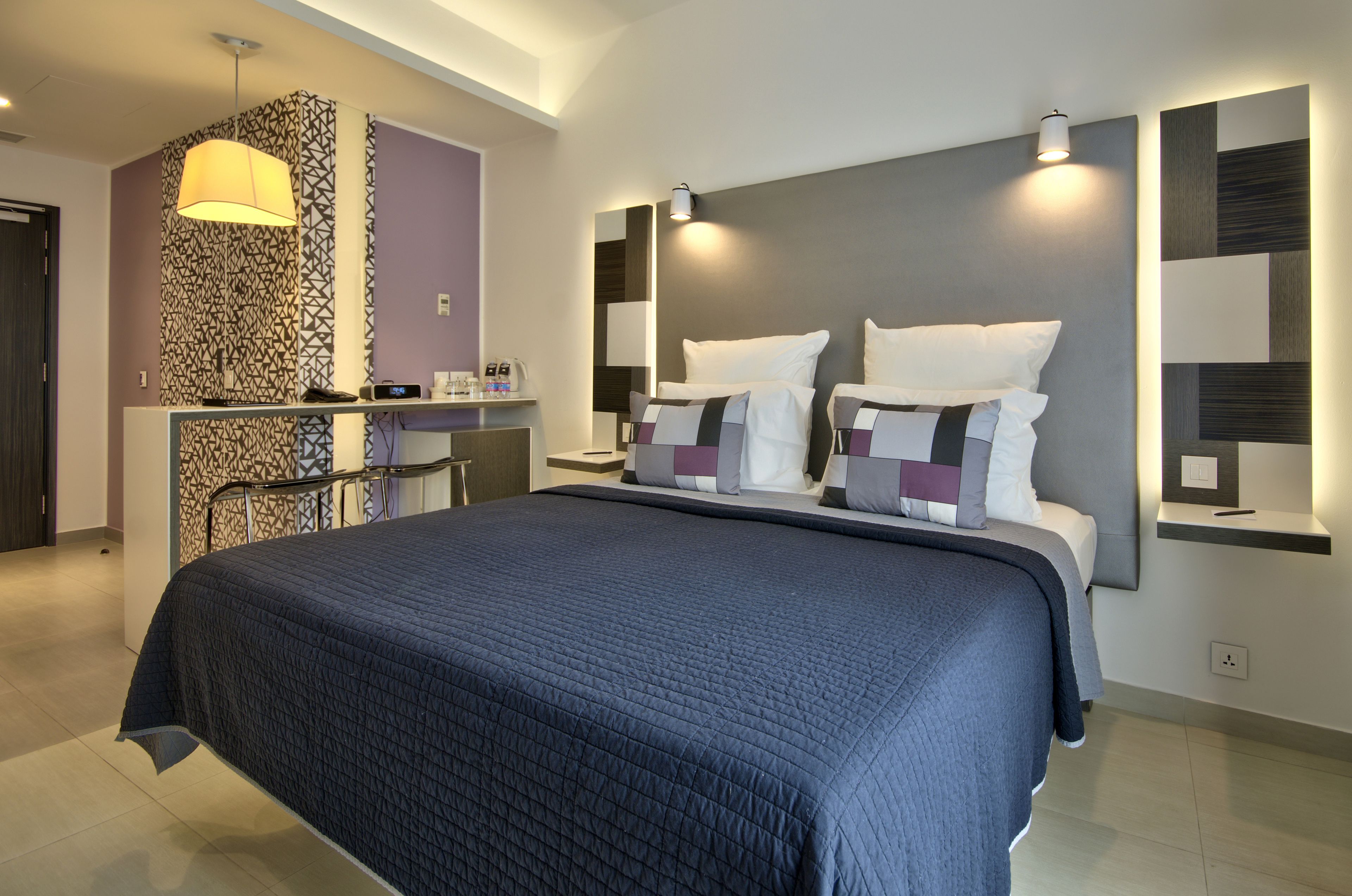 A modern hotel room with a large bed, a desk, and a patterned wall.