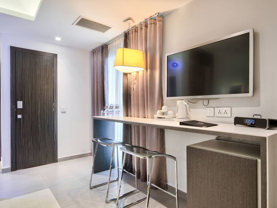 Habitación de hotel moderna con un escritorio, dos taburetes tipo bar, un televisor, una lámpara y un reloj.