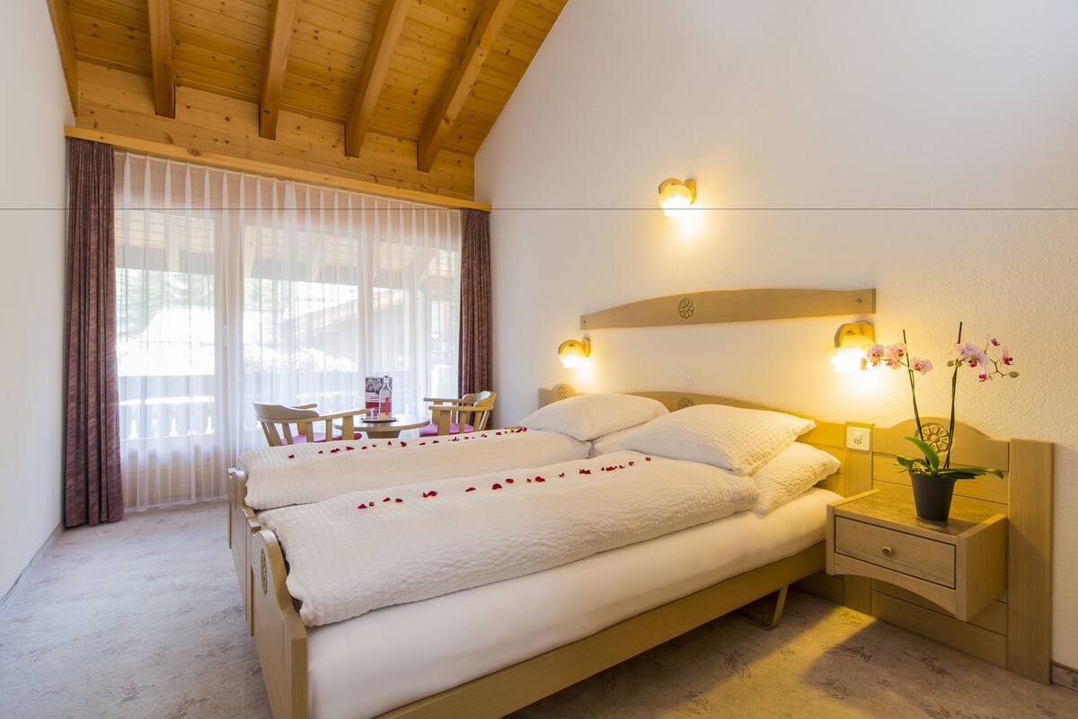 A hotel room with two beds, a wooden ceiling, a small table with chairs, and a nightstand with a plant.
