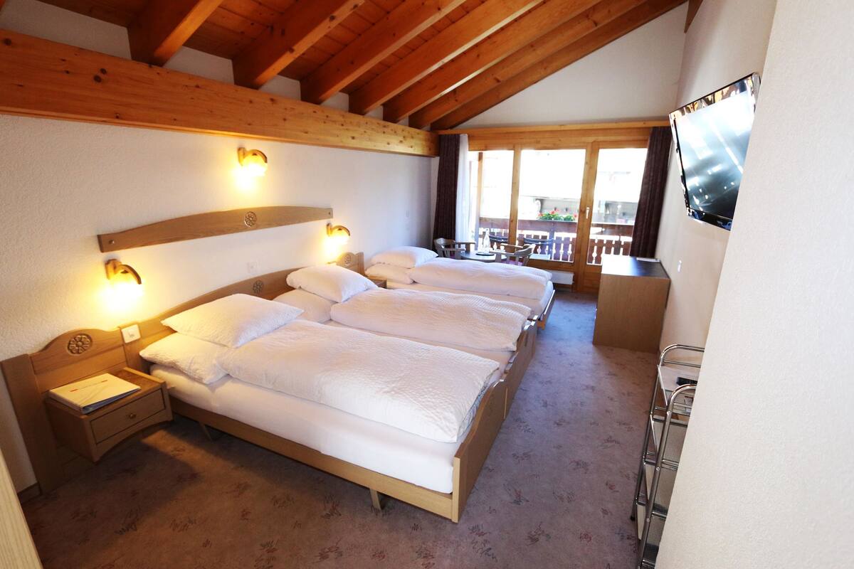 A hotel room with a wooden ceiling, a large bed with white bedding, a nightstand, a TV mounted on the wall, and a balcony with a view.