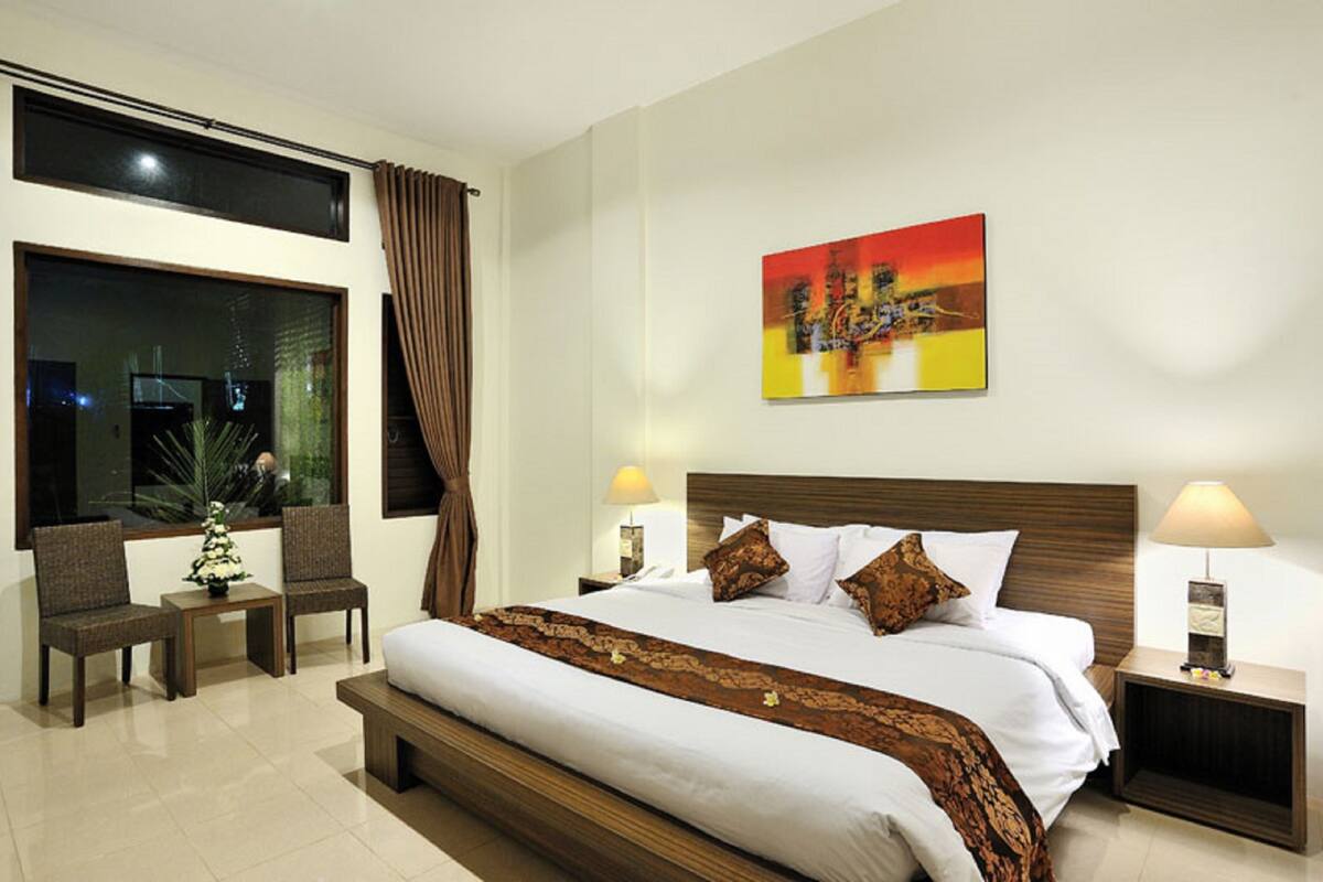 A hotel room with a large bed, two bedside tables with lamps, a window with curtains, a small table with a plant, and a painting on the wall.